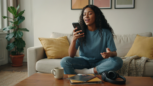 A young woman sings into her cell phone on a couch in a living room.