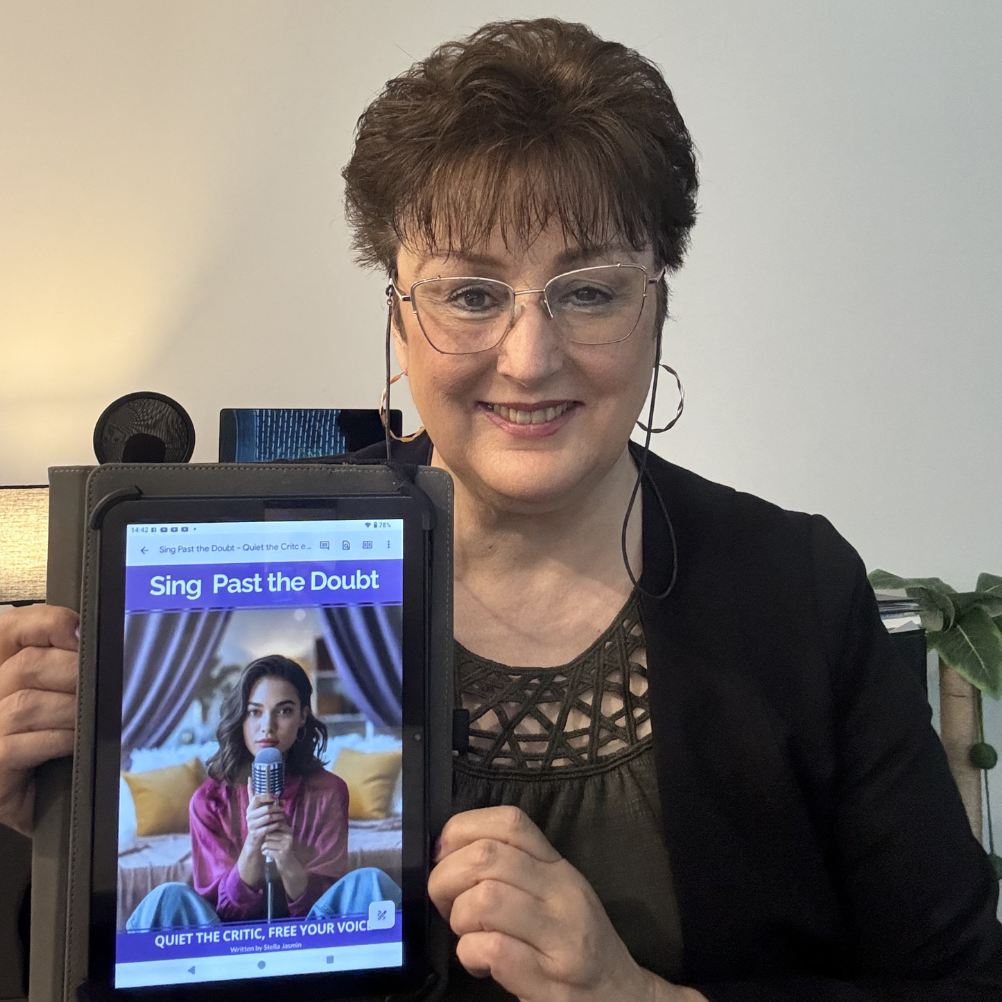 Stella Jasmin holding a tablet displaying the e-book, 'Sing Past The Doubt - Quiet The Critic, Free Your Voice' in an indoor setting.