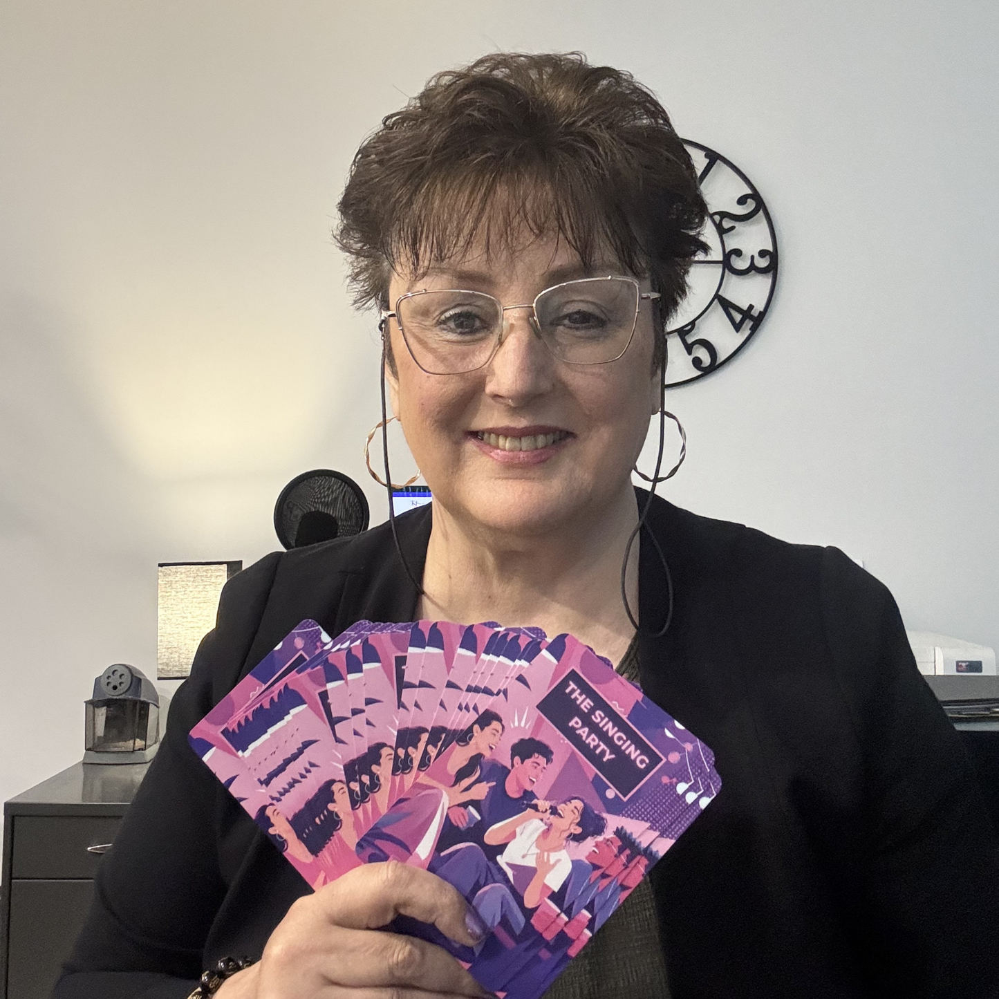 Stella Jasmin holding a purple card game titled 'The Singing Party' in an indoor setting.