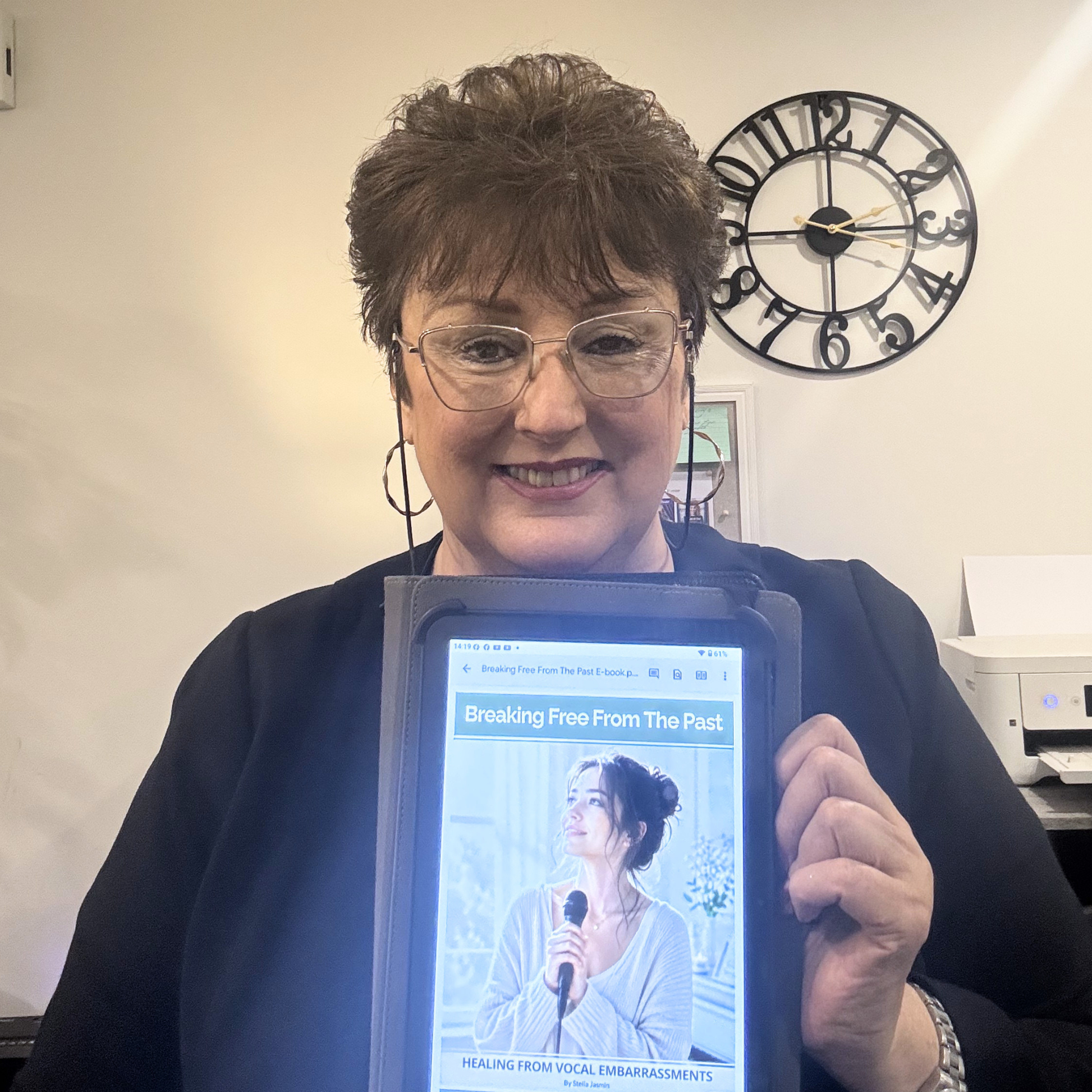 Stella Jasmin holding a tablet with a screen displaying an image of a woman and text for 'Breaking Free From The Past - Healing From Vocal Embarrassments' e-book.